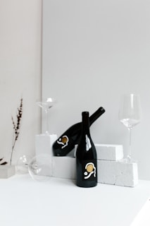 Two black wine bottles are placed on white bricks. One bottle is standing upright while the other is lying on its side. Two empty wine glasses are positioned nearby, and there is a dried plant stem in the background.