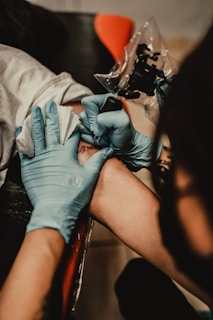A tattoo artist wearing blue gloves is in the process of giving a tattoo on a person's arm. The tattoo machine is visible as the artist works on the design, while the client's arm rests on a surface.