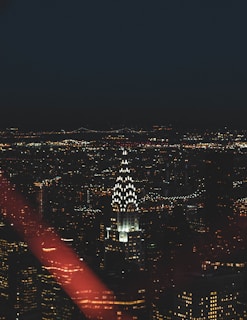 A nighttime cityscape featuring a brightly lit skyscraper with a distinctive art-deco crown surrounded by a sea of city lights. The skyline is dotted with various buildings, and distant bridges are faintly visible against the night sky.