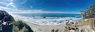 A panoramic view of a beach with a clear blue sky and gentle waves lapping at the shore. The scene features a wooden stairway leading down to the sandy beach surrounded by lush green vegetation on the cliffs. A few people are seen walking along the beach, while others are standing near the cliffs taking photos.