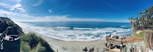 A panoramic view of a beach with a clear blue sky and gentle waves lapping at the shore. The scene features a wooden stairway leading down to the sandy beach surrounded by lush green vegetation on the cliffs. A few people are seen walking along the beach, while others are standing near the cliffs taking photos.