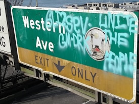 A green and yellow road sign for Western Avenue Exit Only is covered in graffiti with phrases like 'UNITE THE PEOPLE'. A small emblem featuring a stylized cityscape with a space needle is present. The setting appears to be outdoors near a waterfront.