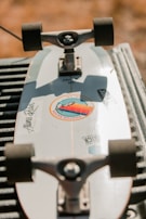 A skateboard is lying upside down on a surface, with its trucks and wheels visible. The skateboard deck is white with stickers on it, including a circular sticker with colored design and text. The background is blurred, giving an outdoor setting and warm lighting.