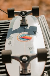 A skateboard is lying upside down on a surface, with its trucks and wheels visible. The skateboard deck is white with stickers on it, including a circular sticker with colored design and text. The background is blurred, giving an outdoor setting and warm lighting.