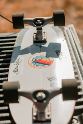 A skateboard is lying upside down on a surface, with its trucks and wheels visible. The skateboard deck is white with stickers on it, including a circular sticker with colored design and text. The background is blurred, giving an outdoor setting and warm lighting.