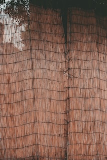 A close-up view of a woven bamboo or reed mat with thin, vertical lines creating a grid-like pattern. The texture appears natural with a slight variation in the hue of the material.