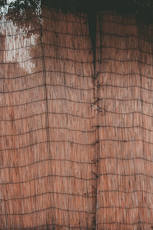 A close-up view of a woven bamboo or reed mat with thin, vertical lines creating a grid-like pattern. The texture appears natural with a slight variation in the hue of the material.