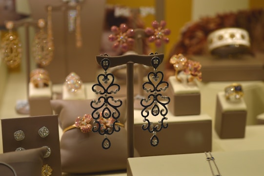 A display case filled with various types of jewelry including ornate earrings, rings, and bracelets, all set on beige display stands. The background features blurred images of floral designs and metallic patterns, contributing to an elegant and opulent atmosphere.