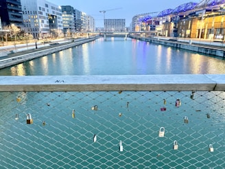 A waterway bordered by modern buildings and crossed by a bridge with numerous locks attached to its wire fencing. The locks are varying in size and color, symbolizing romantic gestures. The scene is illuminated by city lights reflecting on the water, and a crane is visible in the distance, indicating ongoing construction.