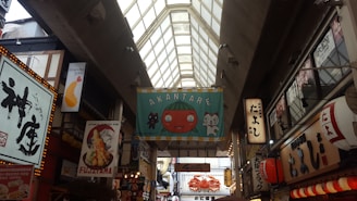 A bustling indoor market with a high glass ceiling, featuring various colorful and illuminated signs in Japanese. A prominent banner in the center displays a cartoon tomato and cat with the text 'AKANTARE'. The scene is filled with bright lights and shops, including a tempura restaurant highlighted by a shrimp illustration.