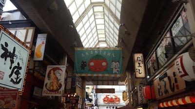 A bustling indoor market with a high glass ceiling, featuring various colorful and illuminated signs in Japanese. A prominent banner in the center displays a cartoon tomato and cat with the text 'AKANTARE'. The scene is filled with bright lights and shops, including a tempura restaurant highlighted by a shrimp illustration.