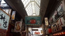 A bustling indoor market with a high glass ceiling, featuring various colorful and illuminated signs in Japanese. A prominent banner in the center displays a cartoon tomato and cat with the text 'AKANTARE'. The scene is filled with bright lights and shops, including a tempura restaurant highlighted by a shrimp illustration.