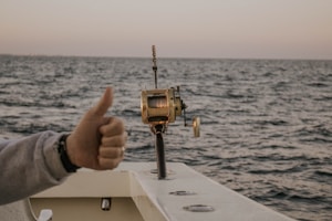 A boat fishing trip on the open sea with a fishing reel mounted on the side of the boat. The ocean stretches to the horizon under a clear sky, and a person gives a thumbs-up gesture, indicating approval or success.