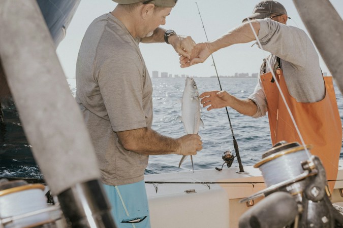 Two anglers on a boat handing off a small silver fish, one in orange bib overalls, with rods and a spinning reel in front and a city skyline across the water