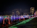 A nighttime scene showcasing a brightly lit park with colorful light displays. Rows of lights in arches span over a walkway adjacent to a body of water, with reflections visible on the surface. People walk along the path, enjoying the festive atmosphere. Buildings with illuminated windows are visible in the background, and trees line the edge of the park.