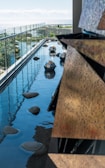 A modern outdoor water feature with smooth stones and reflective metallic spheres placed on a narrow pool next to a glass railing. The ocean is visible in the background, creating a serene and harmonious environment.