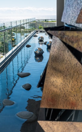 A modern outdoor water feature with smooth stones and reflective metallic spheres placed on a narrow pool next to a glass railing. The ocean is visible in the background, creating a serene and harmonious environment.