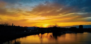 A vibrant sunset paints the sky with shades of orange, yellow, and blue, reflecting on a calm body of water. Trees and silhouettes of buildings stand against the colorful sky, adding depth to the serene landscape.