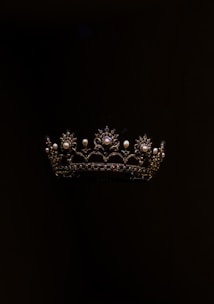 A richly detailed crown featuring intricate patterns and adorned with pearls and gemstones, set against a dark background. The crown's design includes ornate arches and floral motifs, with shimmering elements that stand out against the black backdrop.