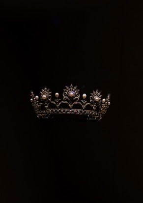 A richly detailed crown featuring intricate patterns and adorned with pearls and gemstones, set against a dark background. The crown's design includes ornate arches and floral motifs, with shimmering elements that stand out against the black backdrop.