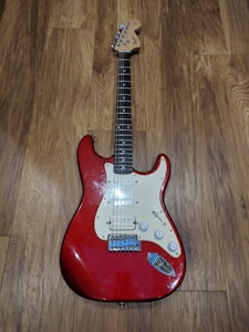 A red electric guitar with a classic Stratocaster shape, featuring a white pickguard, six strings, and control knobs. The guitar is resting on a wooden floor with a visible grain pattern.