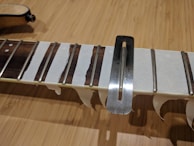 A close-up view of a guitar neck with several frets taped off using masking tape, and a metal fret leveling beam placed across them. The wood grain of the fretboard contrasts with the silver of the frets and the metallic tool.