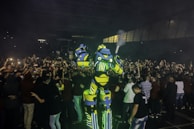 A large and colorful robot costume stands amidst a crowd of people, creating a lively and futuristic atmosphere. The robot is predominantly yellow and blue with glowing green elements. The crowd is taking photos and appears excited and engaged.