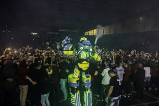 A large and colorful robot costume stands amidst a crowd of people, creating a lively and futuristic atmosphere. The robot is predominantly yellow and blue with glowing green elements. The crowd is taking photos and appears excited and engaged.