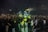 A large and colorful robot costume stands amidst a crowd of people, creating a lively and futuristic atmosphere. The robot is predominantly yellow and blue with glowing green elements. The crowd is taking photos and appears excited and engaged.