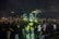 A large and colorful robot costume stands amidst a crowd of people, creating a lively and futuristic atmosphere. The robot is predominantly yellow and blue with glowing green elements. The crowd is taking photos and appears excited and engaged.