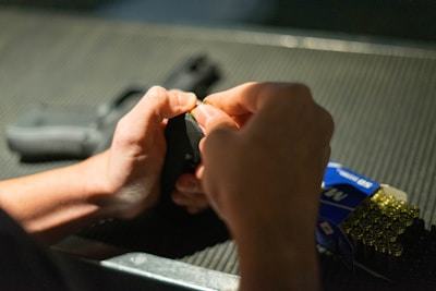 Hands are loading bullets into a firearm magazine. A partially visible handgun rests in the background on a textured surface. To the right, a box of ammunition is open, displaying several rows of bullets.