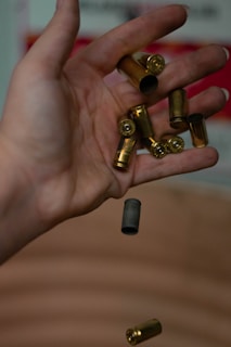 A hand is holding several empty bullet casings, with some falling through the air. The casings are metallic and have a cylindrical shape, with the primer end visible on some of them.