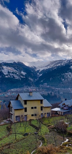 A serene mountainous landscape features a two-story house with a pale yellow facade and a grey roof, surrounded by a lush grassy area with scattered trees. In the distance, snow-capped mountains rise under a dramatic sky filled with large, billowing clouds. A neighboring house is positioned slightly downhill, and the view extends to a valley with more buildings visible.