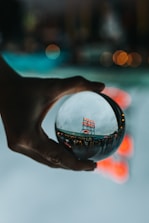 A hand is holding a crystal ball, through which a busy public market sign is viewed in a distorted, artistic manner. The background is blurred with bokeh lights, creating a dreamy and reflective ambiance.