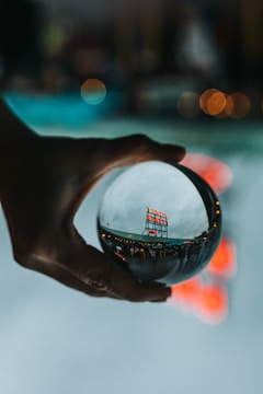 A hand is holding a crystal ball, through which a busy public market sign is viewed in a distorted, artistic manner. The background is blurred with bokeh lights, creating a dreamy and reflective ambiance.