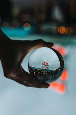 A hand is holding a crystal ball, through which a busy public market sign is viewed in a distorted, artistic manner. The background is blurred with bokeh lights, creating a dreamy and reflective ambiance.