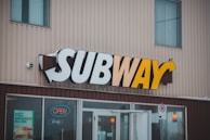 A storefront features the recognizable 'SUBWAY' sign with bold letters that transition from white to yellow. Below the sign, the entrance has glass doors, one of which has an 'open' sign illuminated in red. To the left of the entrance, there's a promotional poster advertising a $2.99 breakfast offer. The building's exterior is made of vertical beige siding panels.
