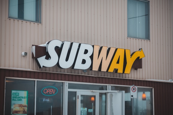 A storefront features the recognizable 'SUBWAY' sign with bold letters that transition from white to yellow. Below the sign, the entrance has glass doors, one of which has an 'open' sign illuminated in red. To the left of the entrance, there's a promotional poster advertising a $2.99 breakfast offer. The building's exterior is made of vertical beige siding panels.