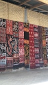 A series of intricately patterned textiles are hanging vertically against a woven bamboo wall. The textiles feature a variety of traditional motifs including animals and geometric patterns in rich colors such as red, black, and white.