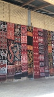 A series of intricately patterned textiles are hanging vertically against a woven bamboo wall. The textiles feature a variety of traditional motifs including animals and geometric patterns in rich colors such as red, black, and white.