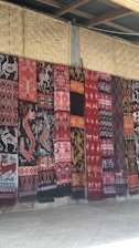 A series of intricately patterned textiles are hanging vertically against a woven bamboo wall. The textiles feature a variety of traditional motifs including animals and geometric patterns in rich colors such as red, black, and white.