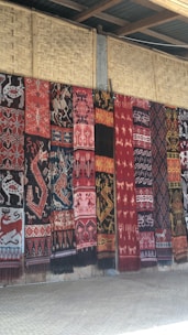 A series of intricately patterned textiles are hanging vertically against a woven bamboo wall. The textiles feature a variety of traditional motifs including animals and geometric patterns in rich colors such as red, black, and white.