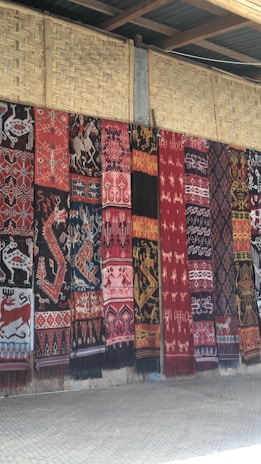 A series of intricately patterned textiles are hanging vertically against a woven bamboo wall. The textiles feature a variety of traditional motifs including animals and geometric patterns in rich colors such as red, black, and white.