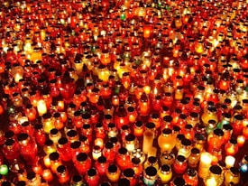 A multitude of lit candles placed closely together, creating a warm, glowing sea of light. The candles vary in size and shape, with many housed in red glass containers, adding a rich, vibrant hue to the scene.