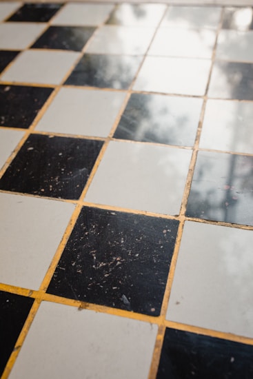A checkered floor pattern composed of alternating black and white tiles, with a slightly worn and reflective surface. The grout between the tiles is yellowish in color, contributing to a nostalgic or vintage appearance.