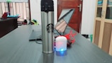 A stainless steel tumbler with a recognizable logo is placed on a wooden desk next to a laptop. White earphones with a charging case and a blue light are visible, along with red cables. The background includes a window, a closed wooden door, shelves with items, and a partially visible backpack.