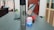 A stainless steel tumbler with a recognizable logo is placed on a wooden desk next to a laptop. White earphones with a charging case and a blue light are visible, along with red cables. The background includes a window, a closed wooden door, shelves with items, and a partially visible backpack.