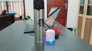 A stainless steel tumbler with a recognizable logo is placed on a wooden desk next to a laptop. White earphones with a charging case and a blue light are visible, along with red cables. The background includes a window, a closed wooden door, shelves with items, and a partially visible backpack.