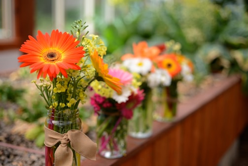 A collection of vibrant flowers displayed in glass jars. The flowers include large orange daisies, surrounded by smaller yellow and white blooms. A brown ribbon is tied around one of the jars, adding a rustic touch. The background features soft-focus greenery, creating a natural and inviting atmosphere.