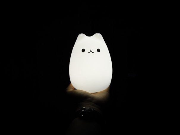 A hand is holding a glowing cat-shaped lamp in a dark environment. The lamp has a cute, minimalistic design with simple facial features, giving it a playful and whimsical appearance. The light from the lamp softly illuminates the hand supporting it.
